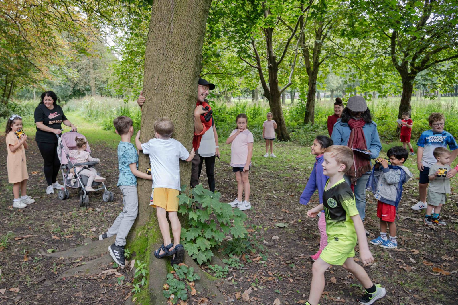 Kids and adults walk excitedly through the park. Paul peers from behind a tree smiling, two kids hug the same tree. Some of the children are holding binoculars.