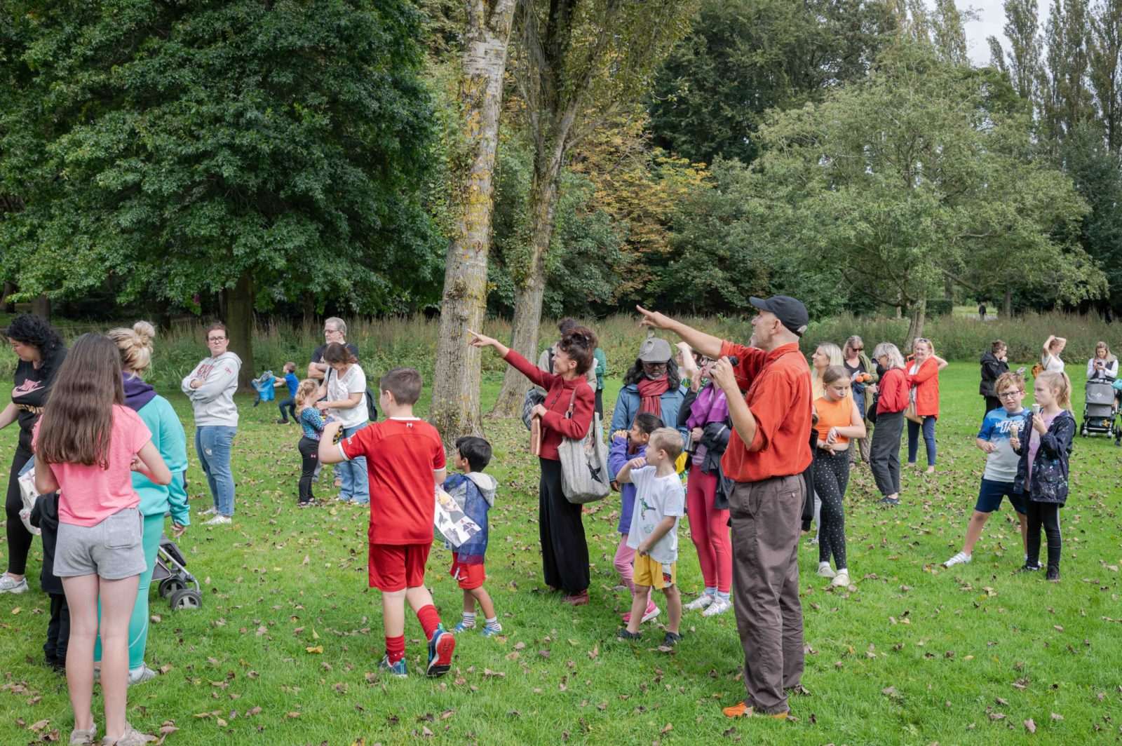 A large group of kids and adults are in the park, Paul is pointing at something in the sky.