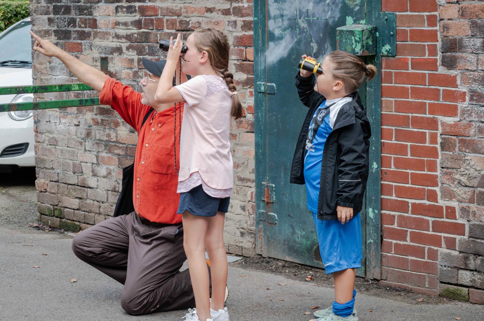 Paul kneels down and points to something in the sky whilst two children next to him look in the same direction with binoculars.
