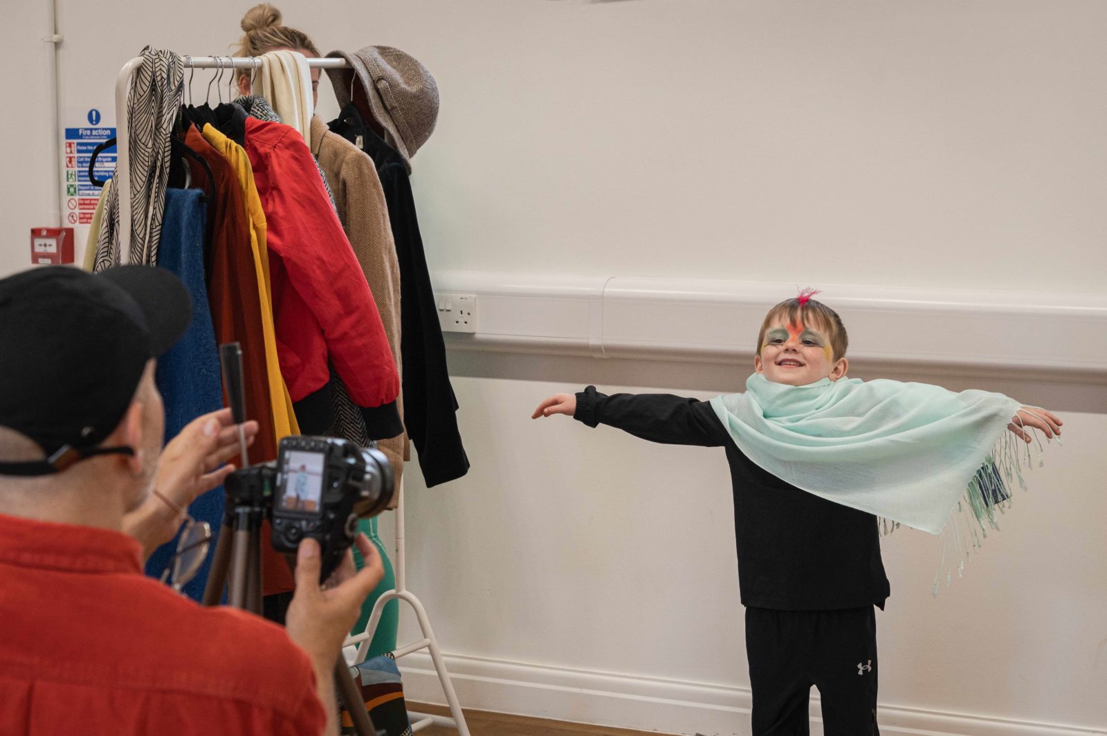 A little boy with blue and red make up on his face, a red feather on his head and a light blue scarf draped over his shoulders smiles at the camera with his arms spread out like wings as Paul takes his photo next to a clothes rail.