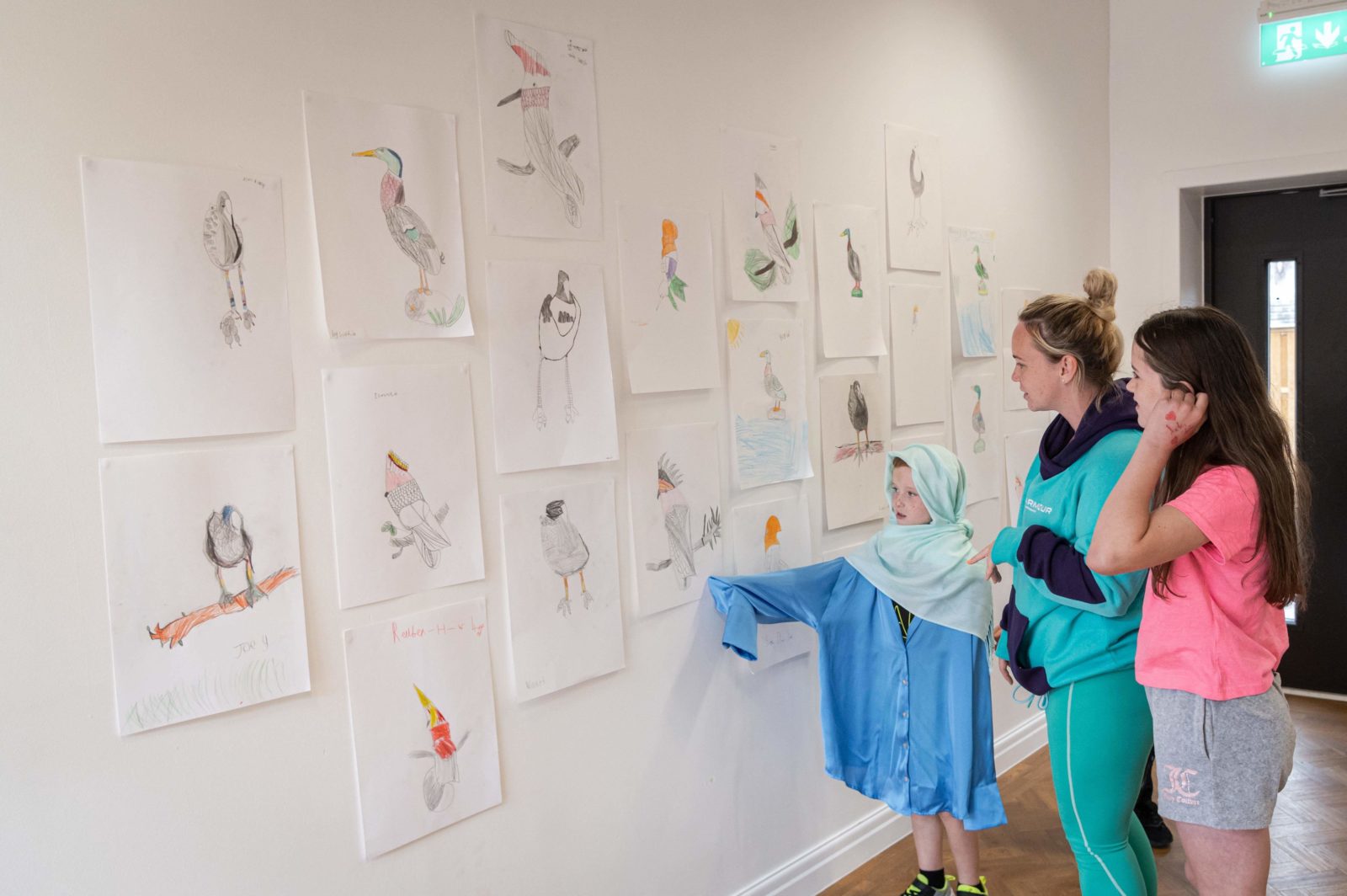 A family is looking at the exhibition of the children's gently referenced bird drawings. The little boy in the photo is pointing at a drawing, wearing an adult's blue shirt that goes down to his knees, and a mint green scarf wrapped around his head and shoulders.