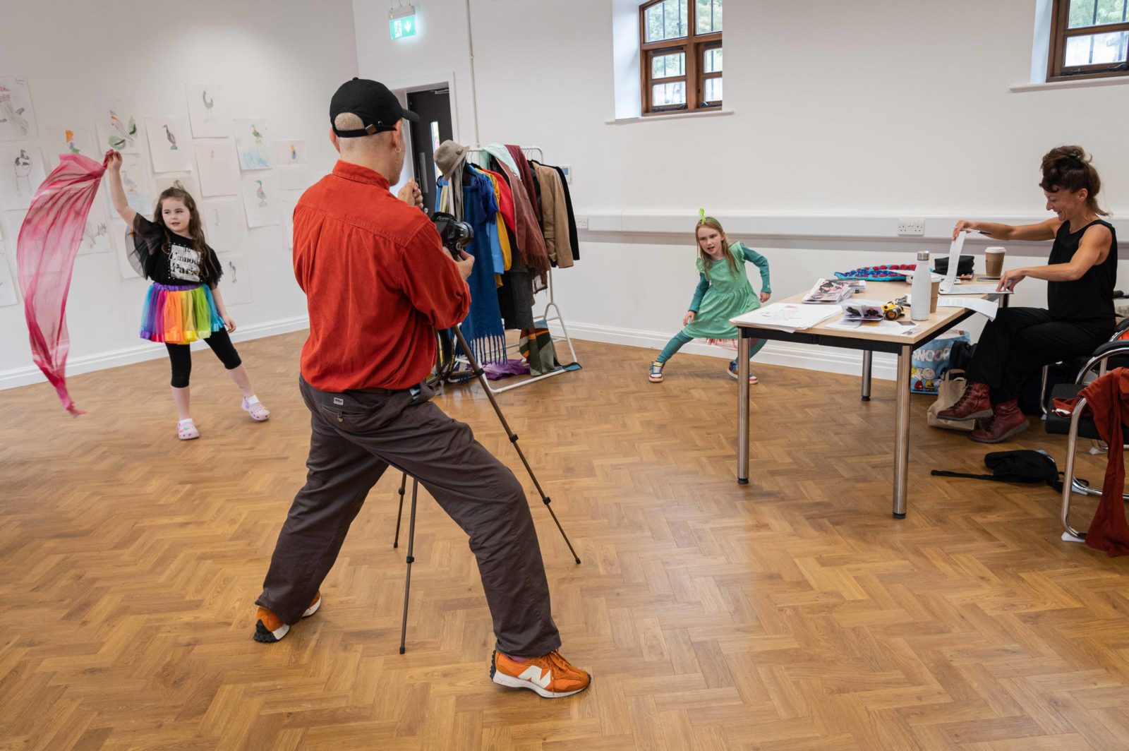 Paul stands behind a tripod taking a little girl's photo, the girl is posing with one leg bent and the other stretched out, leaning to the side, she is wearing all green with a green feather on her head. In the background another little girl dances with a pink silk scarf and a rainbow skirt.