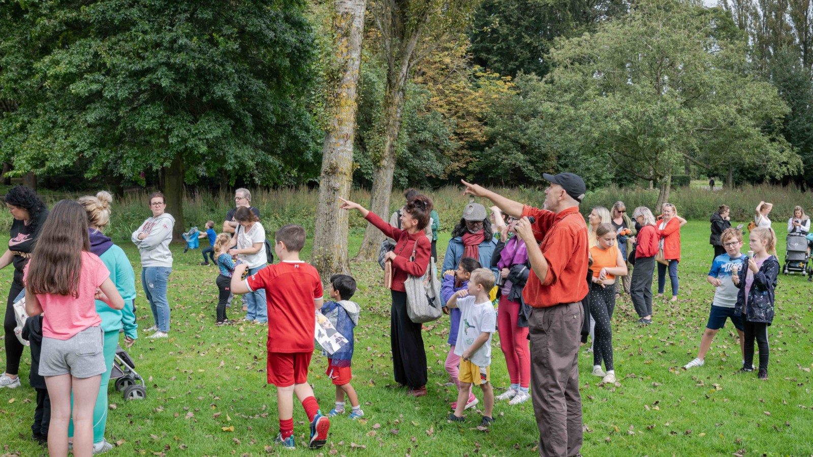 A large group of kids and adults are in the park, Paul is pointing at something in the sky.