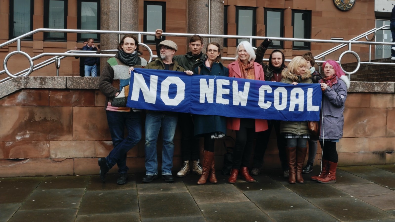 A group of people stand outside court holding a banner that reads 'NO NEW COAL' in white writing on a blue background.