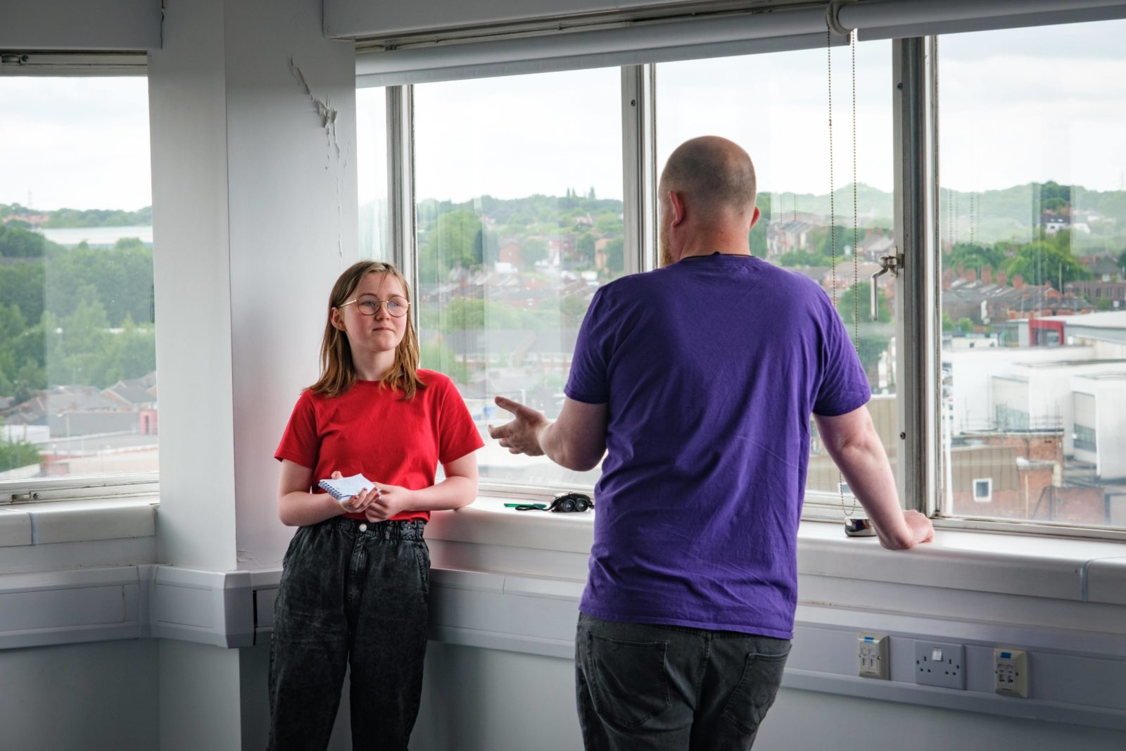 A student holding a notepad talks to a man in front of a row of windows.