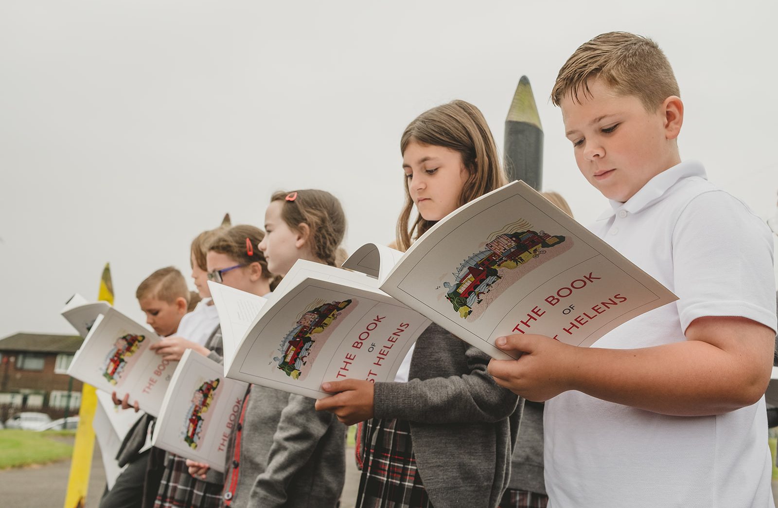 Six children stand in line. They are all wearing school uniforms that include a shirt with either grey trousers or a grey and red pinafore.  They each read The Book Of St Helens