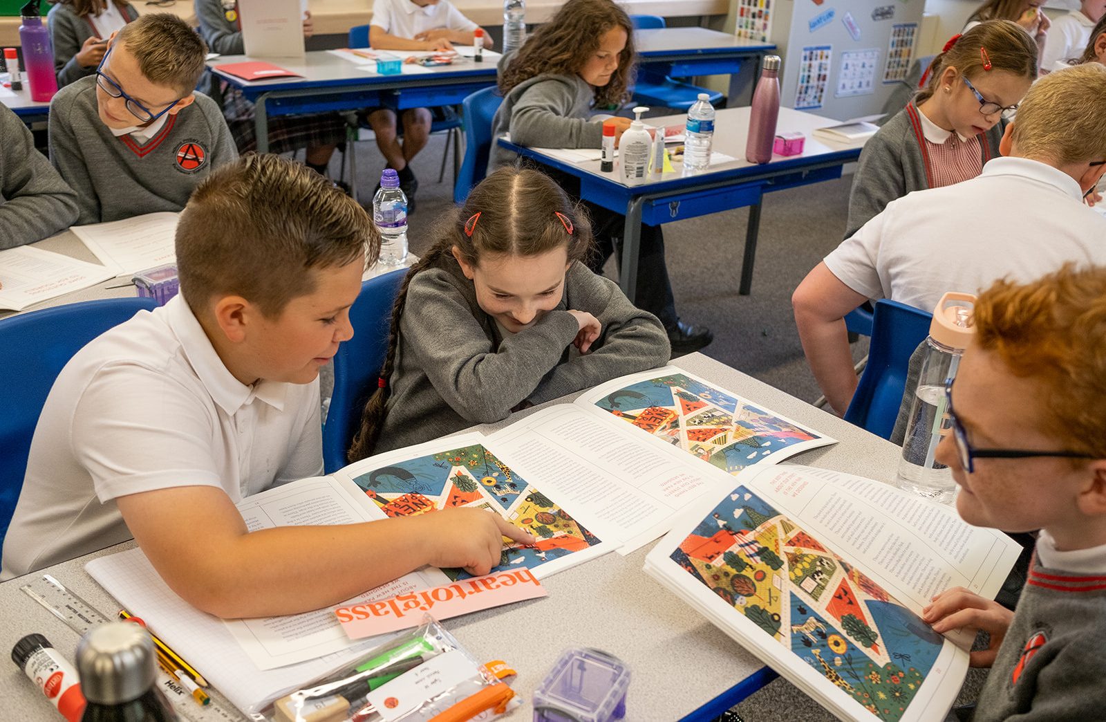 Children are seated at tables in a classroom. They are all reading the book of St Helens and talking to each other about it.