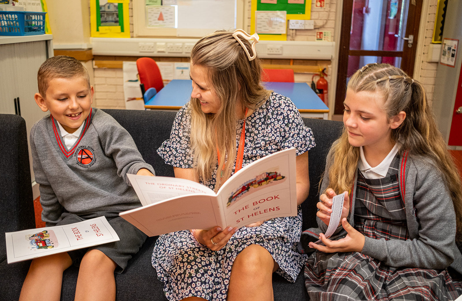 A boy and a girl in school uniforms sit either sit of their teacher on a bench. They are reading the book of St Helens together and are smiling