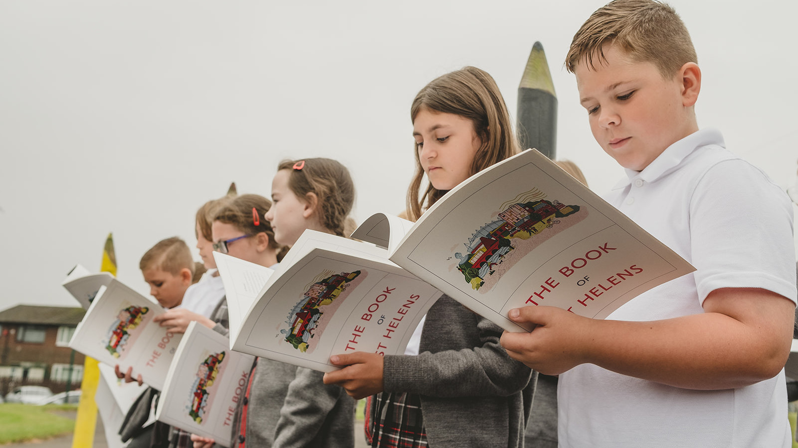 Six children stand in line. They are all wearing school uniforms that include a shirt with either grey trousers or a grey and red pinafore.  They each read The Book Of St Helens