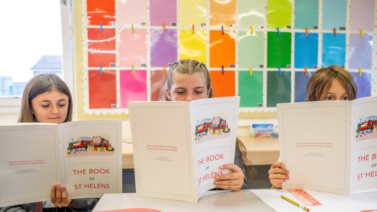 Three school children sit in front of a coloured background reading individual copies of The Book of St Helens