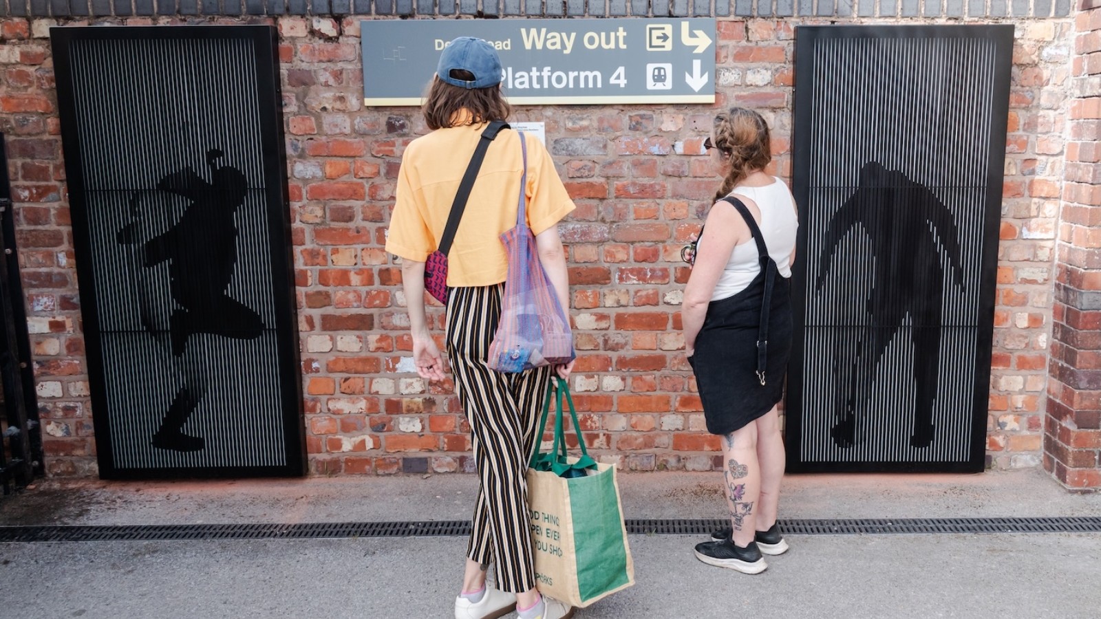 Two people look at an metal artwork on a wall on a station platform. The artwork on the left shows a skipping girl and the one on the right is the shape of a young person dancing