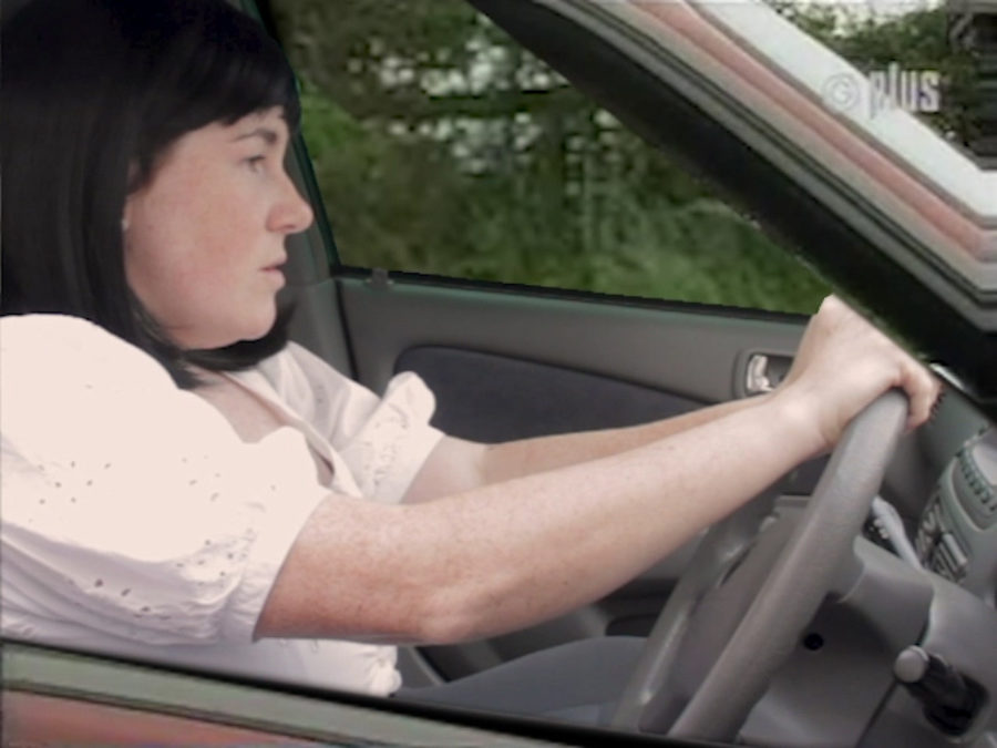 Artist Amy Pennington wears a black shoulder length wig and a white short sleeved top and is driving a car.