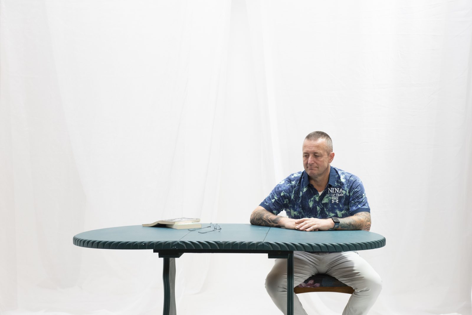 Photograph of a man sat alone at a rippled table, next to a pair of glass and a book placed down on a open on a specific page
