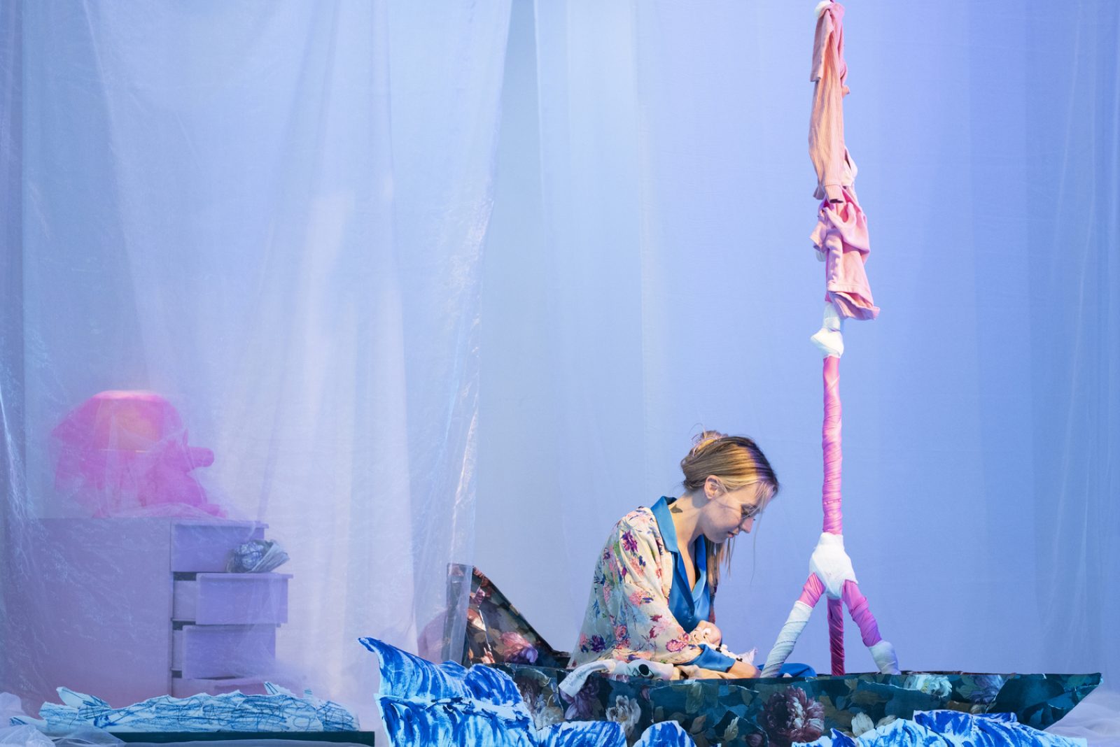 Photograph of a woman wearing pyjamas sat in a paper mache sail boat with plastic sheets hung up around her, and a pink set of drawers behind her