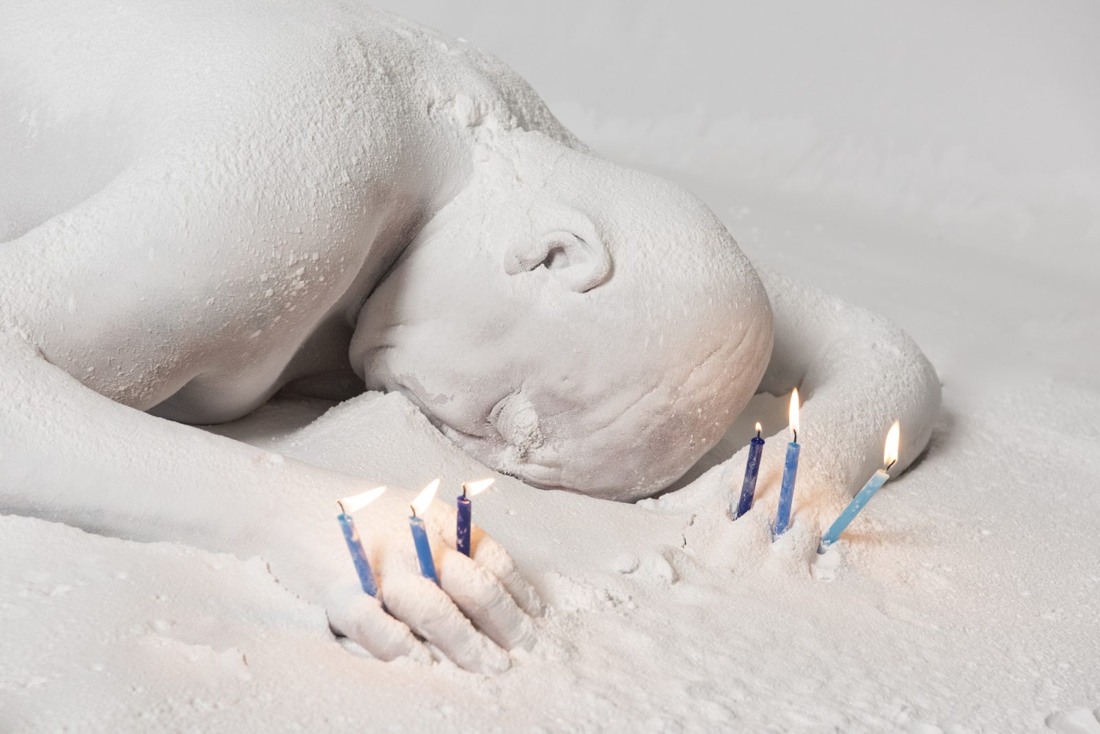 Photograph of a man encased in white sand with three lit birthday candles stuck in between his fingers