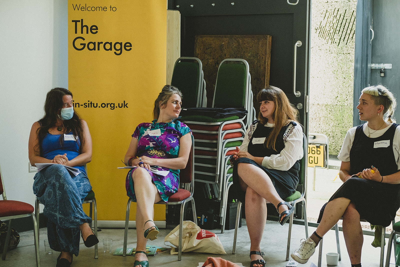 Four women sit on chairs, socially distanced from one another. Behind them is a yellow banner with the words "Welcome to The Garage" and "www.In-Situ.org.uk"