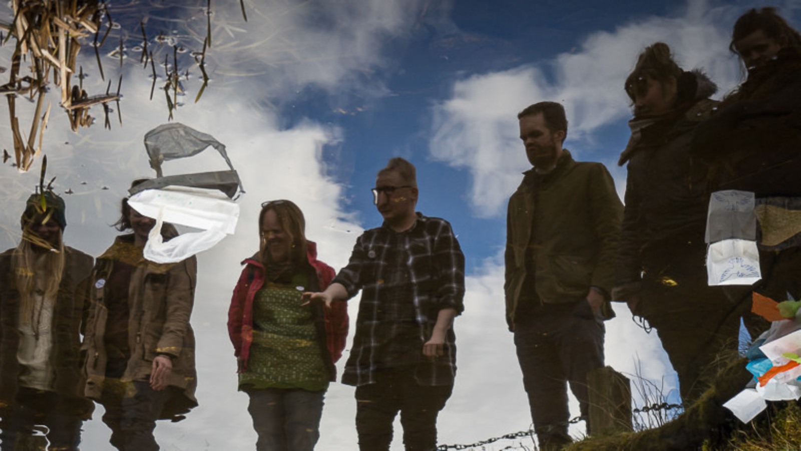 A group of people are photographed looking down at their reflections on a lake that has litter floating on its surface.