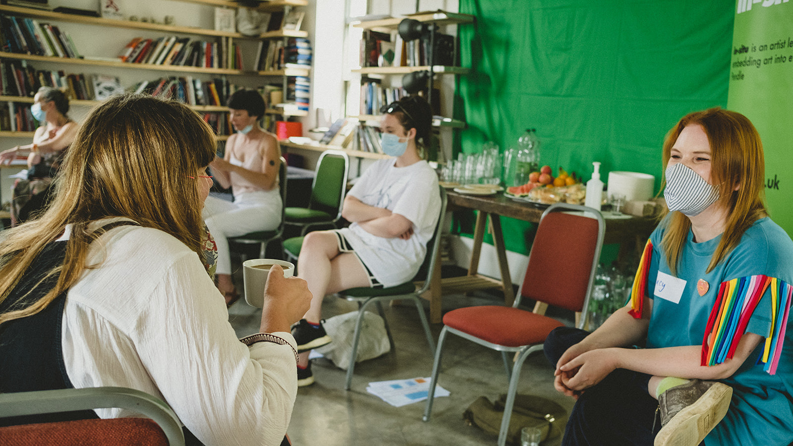 A number of women are seated in a room with vibrant green walls. They are chatting to one another and drinking cups of tea. Some of them are wearing protective face coverings.