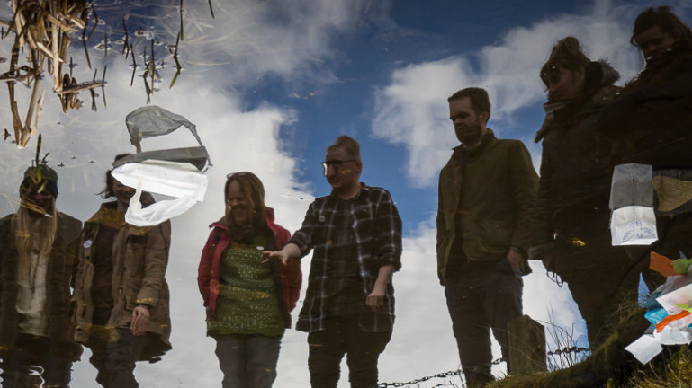A group of people are photographed looking down at their reflections on a lake that has litter floating on its surface.