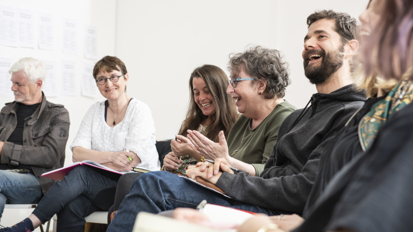 A group of people sit in a row in a white room laughing and talking. They have notepads and books on their laps.