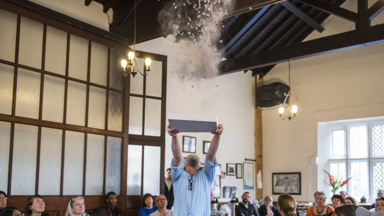 In the middle of a large room with wooden beams and dividers a man stands throwing a cloud of jigsaw pieces and glitter in to the air above his head. An audience look in to the air towards the cloud, some smiling.