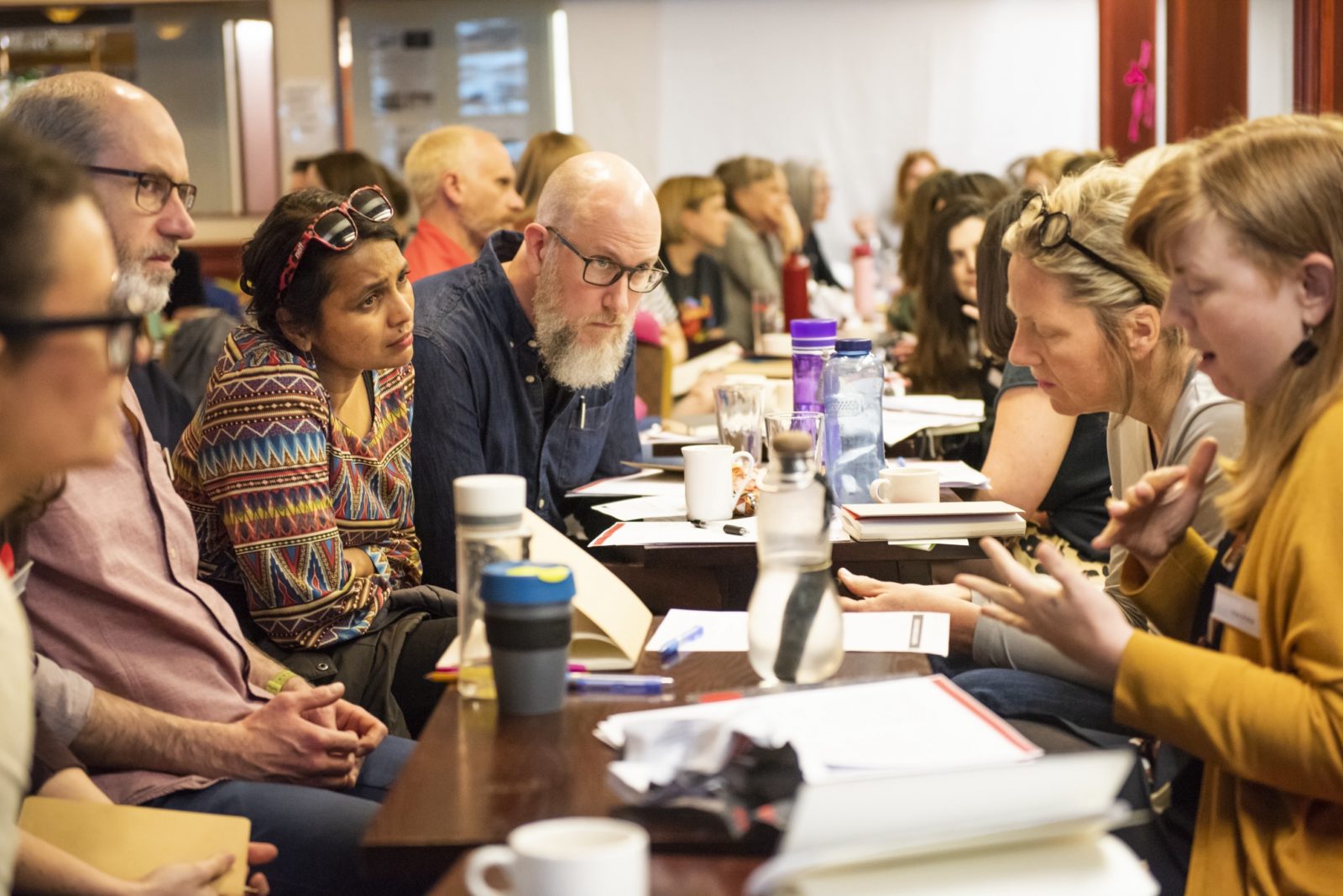 A group of people wearing brightly coloured clothes sit around a table that has papers, water bottles and coffee cups on it. A person with shoulder length light red hair and wearing a yellow cardigan is speaking and gesticulating. The rest of the people appear to be listening to them intensely.