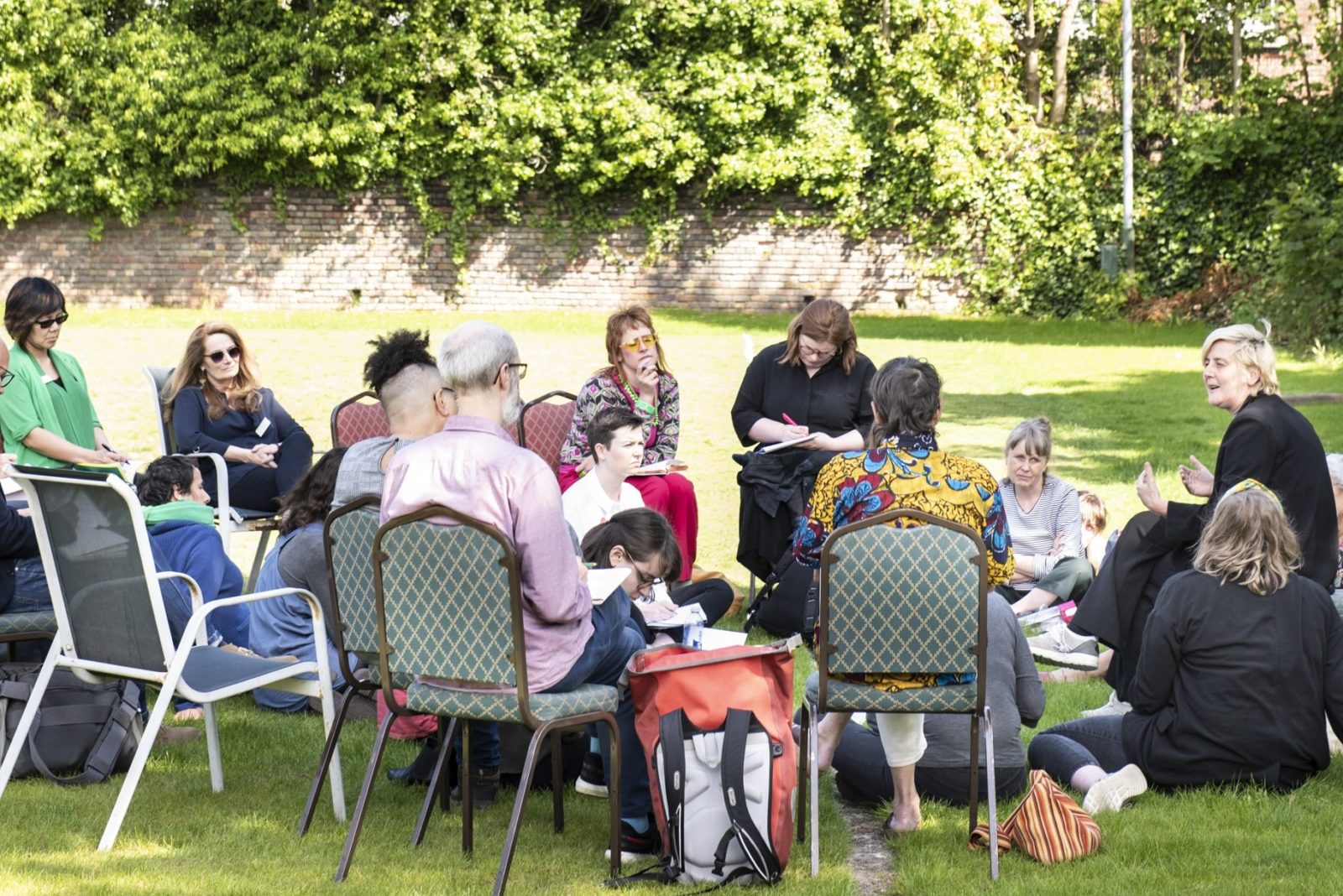 A group of people wearing brightly coloured clothes sit in an informal circle, some are seated on chairs and others on the ground. Some have note pads in their hands and some are writing, some are looking at the speaker, a person with short blonde hair and wearing all black, gesturing with their hands. In the background is a green playing field,  a wall and green foliage.