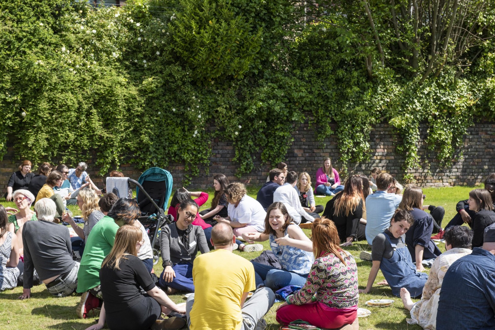 In a large grassy, green space surrounded by trees is busy with people sitting in groups on the grass.