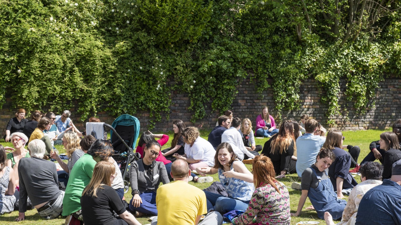 In a large grassy, green space surrounded by trees is busy with people sitting in groups on the grass.