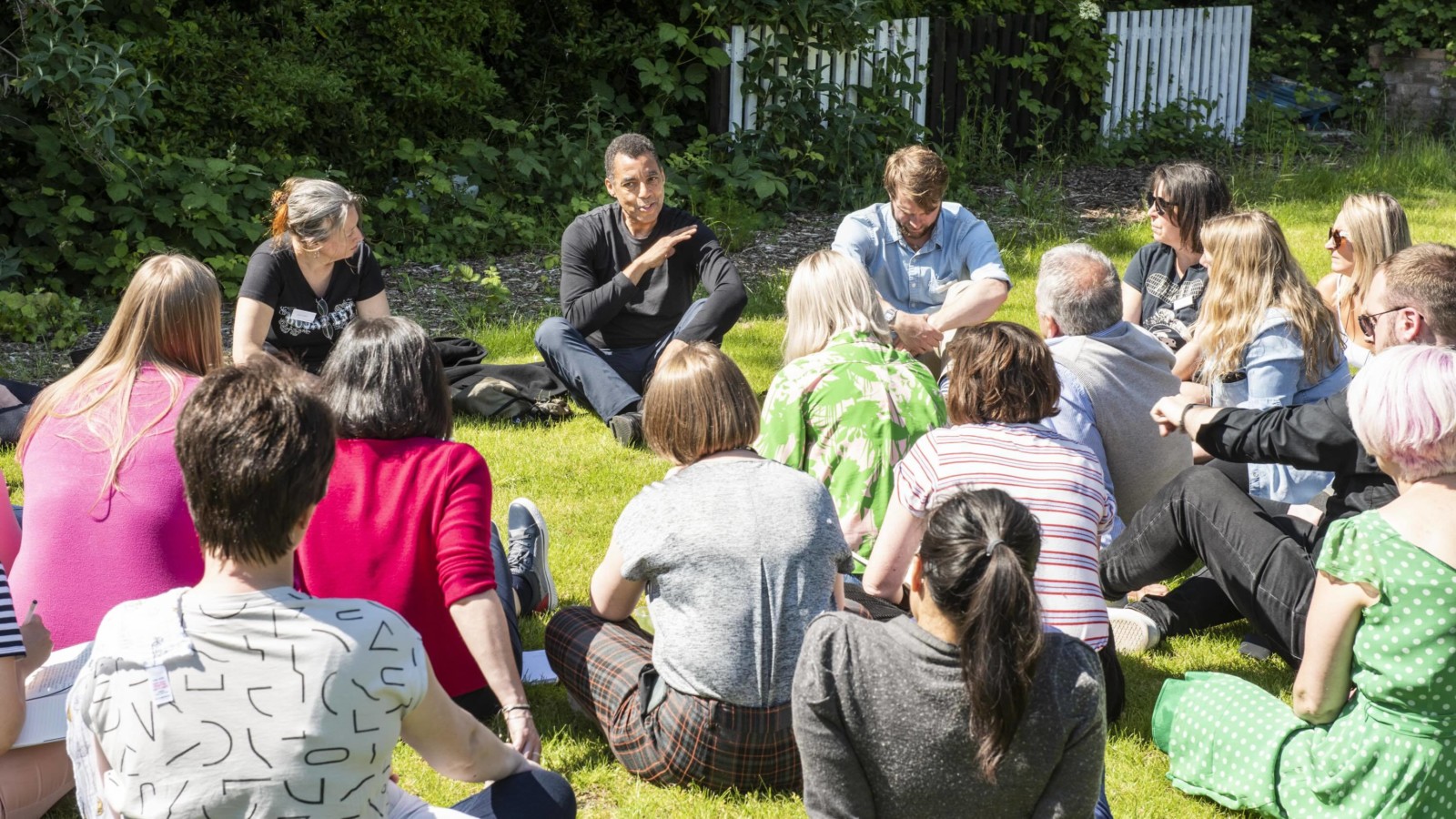 A group of people wearing brightly coloured clothes sit in an informal circle on a patch of grass. They appear to be listening to a speaker, a black man wearing a dark jacket and dark trousers. In the background there is green foliage.