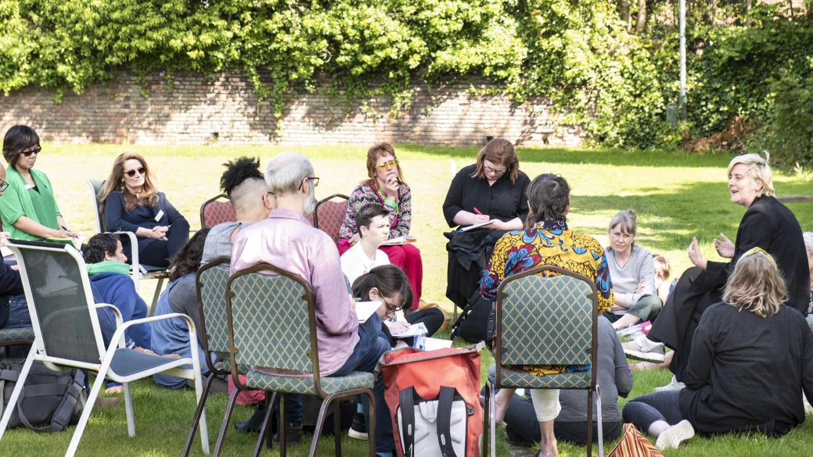 A group of people wearing brightly coloured clothes sit in an informal circle, some are seated on chairs and others on the ground. Some have note pads in their hands and some are writing, some are looking at the speaker, a person with short blonde hair and wearing all black, gesturing with their hands. In the background is a green playing field,  a wall and green foliage.