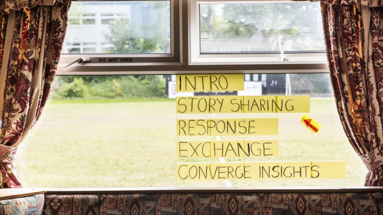 A double glazed window is framed by curtains and a cushioned seat that are patterned in colours of reds, oranges, yellows and pinks. On the window, post its read 'Intro, Story Sharing, Response, Exchange, Converge Insights' - a red arrow points to 'story sharing'. Behind the window there is a bowling green of green grass.