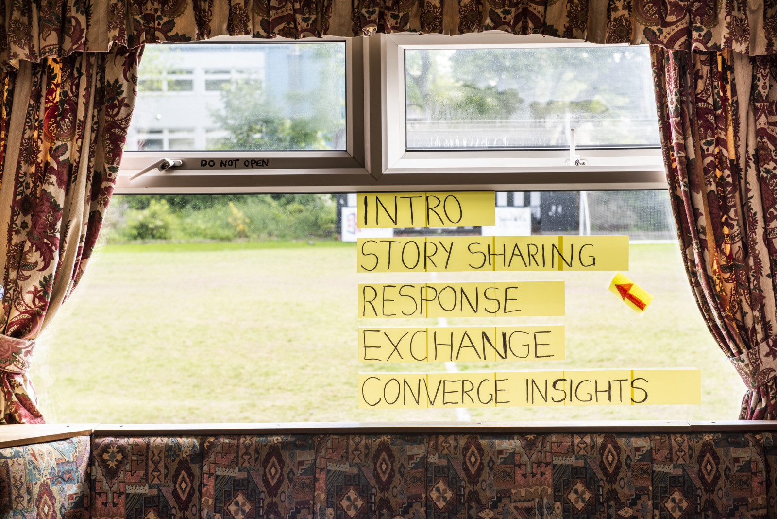 A double glazed window is framed by curtains and a cushioned seat that are patterned in colours of reds, oranges, yellows and pinks. On the window, post its read 'Intro, Story Sharing, Response, Exchange, Converge Insights' - a red arrow points to 'story sharing'. Behind the window there is a bowling green of green grass.