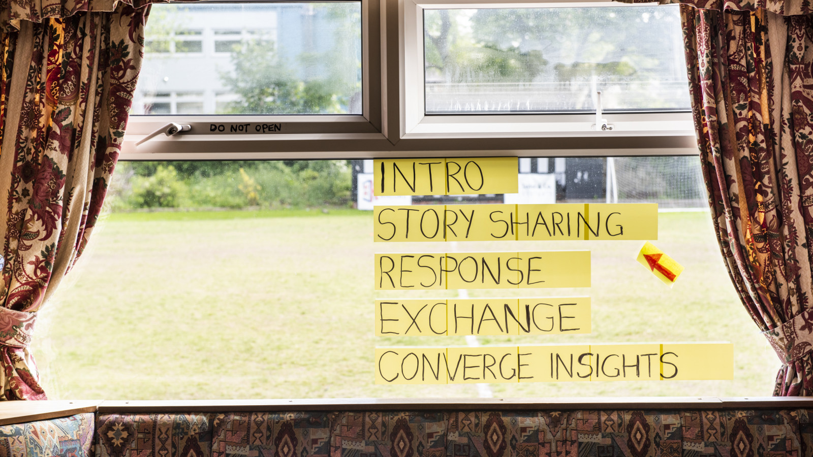 A double glazed window is framed by curtains and a cushioned seat that are patterned in colours of reds, oranges, yellows and pinks. On the window, post its read 'Intro, Story Sharing, Response, Exchange, Converge Insights' - a red arrow points to 'story sharing'. Behind the window there is a bowling green of green grass.