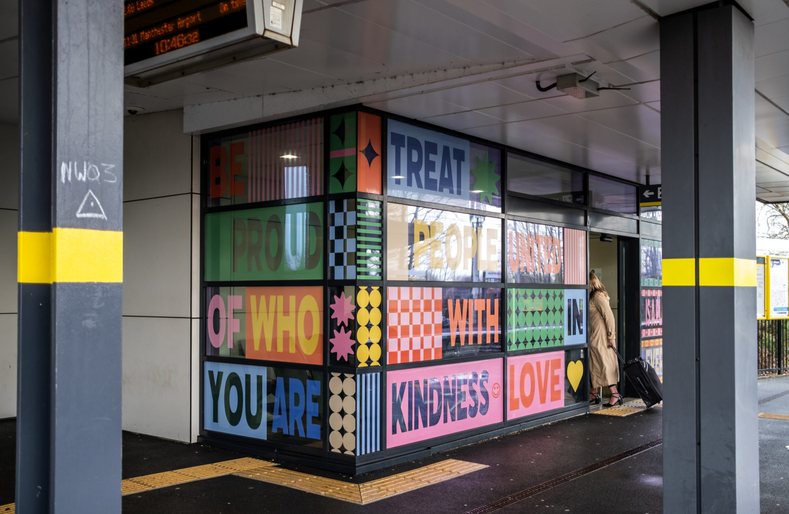 The installation is pictured from slightly further away in between the pillars of the station. A woman walks through the automatic doors with a suitcase.