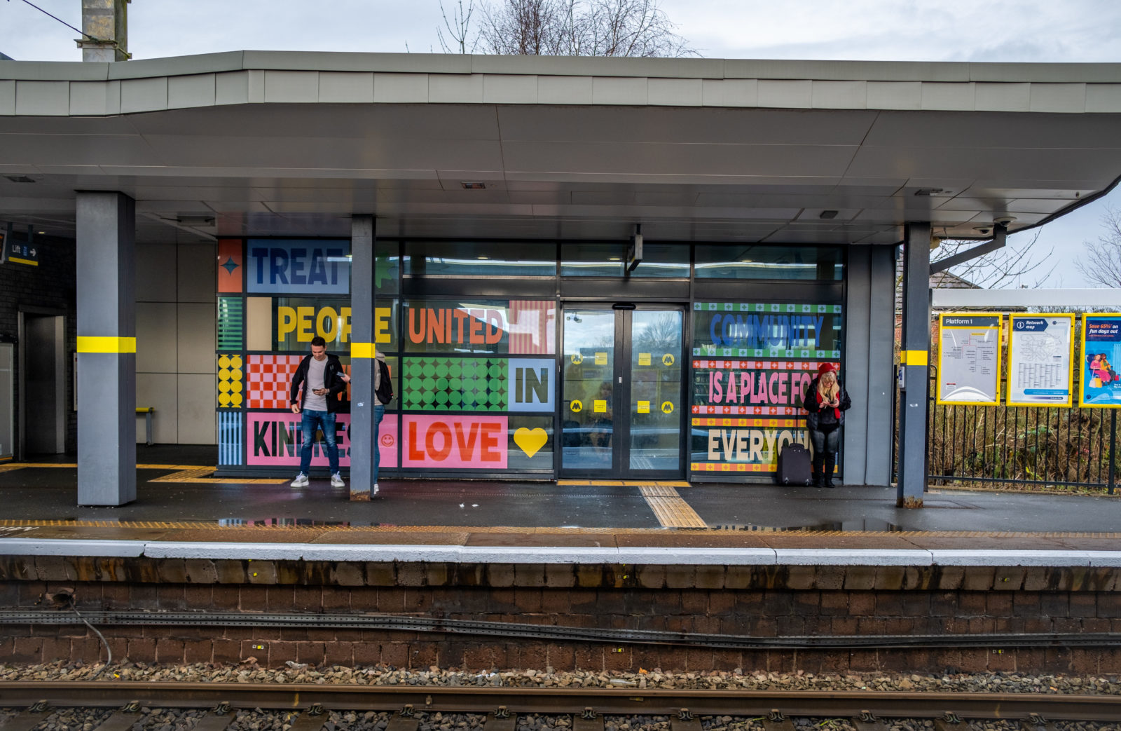 The window installation is photographed from the opposite platform, 3 people stand in front of it waiting for a train.