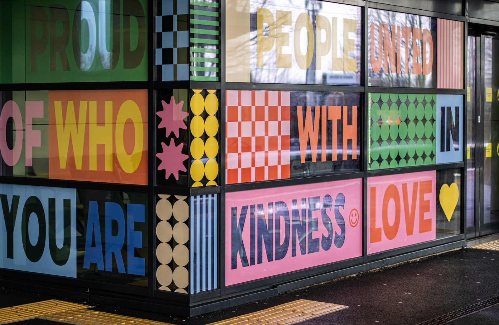 The Queer Eutopia vinyl window installation is photographed up close, it is multi coloured and the words are surrounded by pattern. The words ‘Be Proud of who you are’ ‘Treat People with kindness’ And ‘United in Love’ can be read.