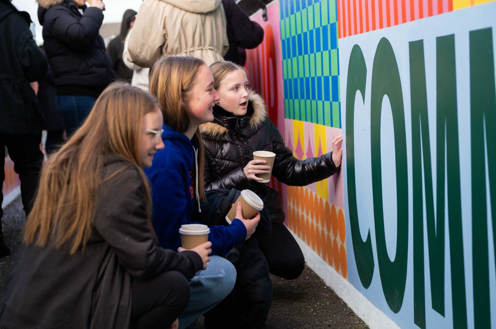 3 young people holding hot drinks look closely at the mural, one is talking with one hand on the wall and the other 2 are smiling.