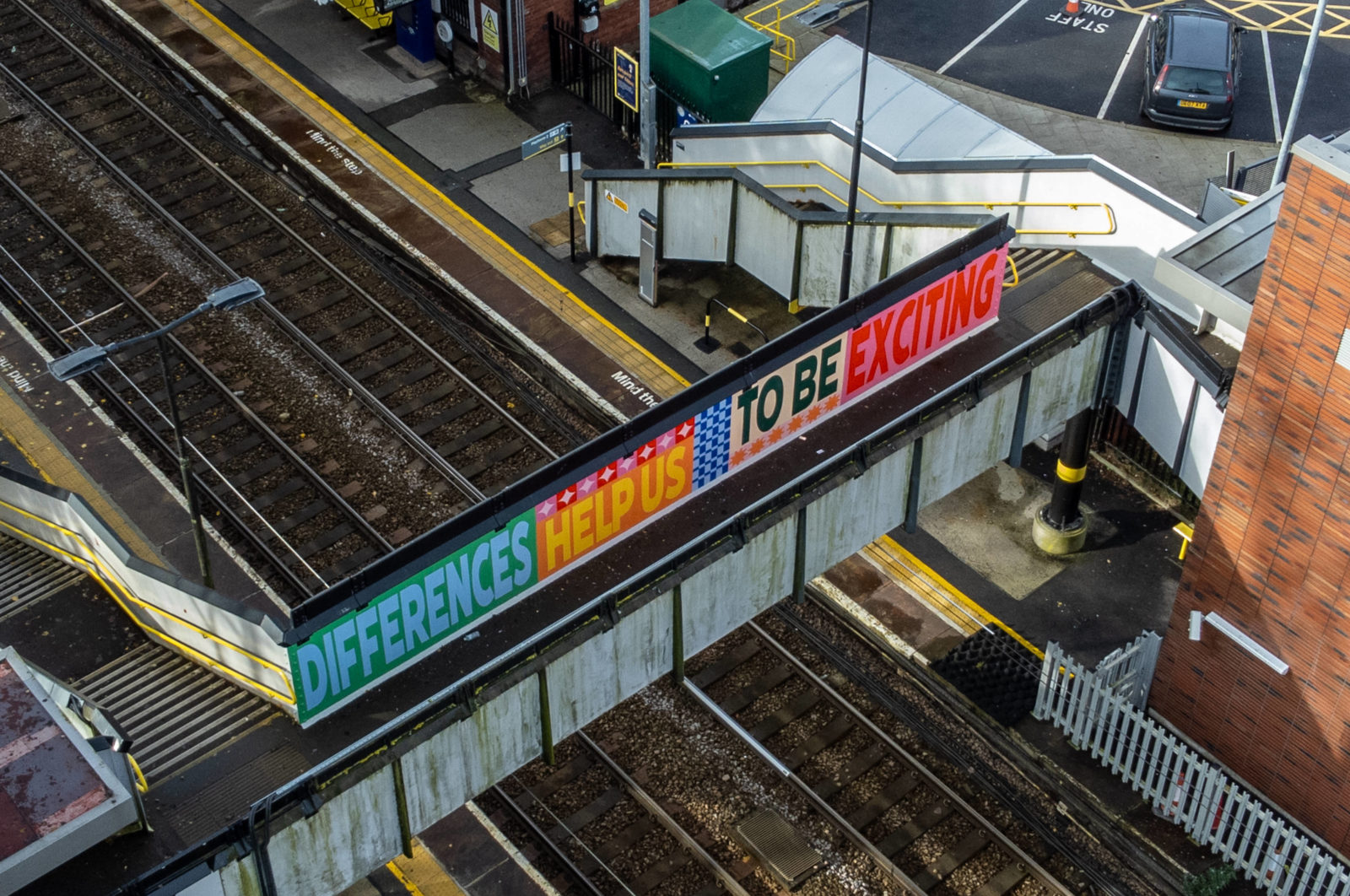 The mural is photographed at an arial view, it reads 'Differences help us to be exciting', surrounded by pattern and is painted on a train station bridge.