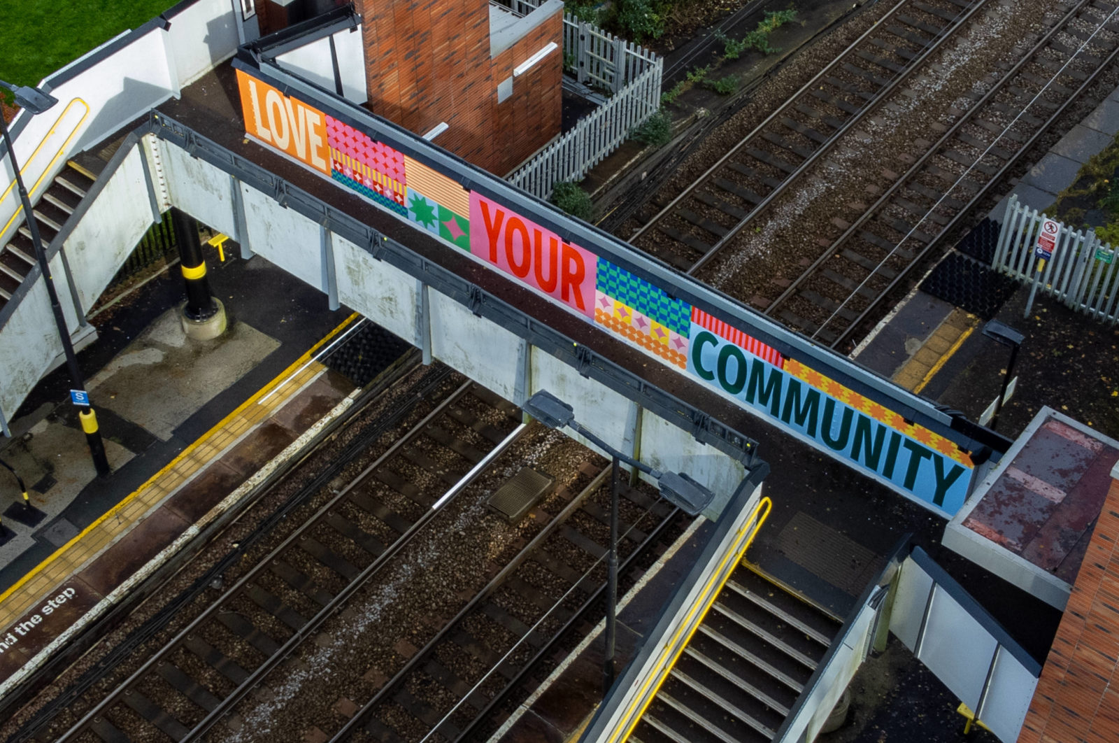 The mural is photographed at an arial view, it reads 'Love Your Community', surrounded by pattern and is painted on a train station bridge.