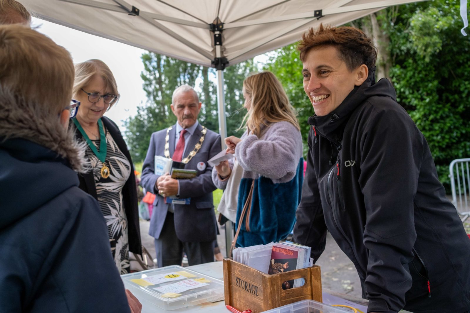 Leo is photographed smiling in conversation with a flower show attendee from behind a table with postcards on it.