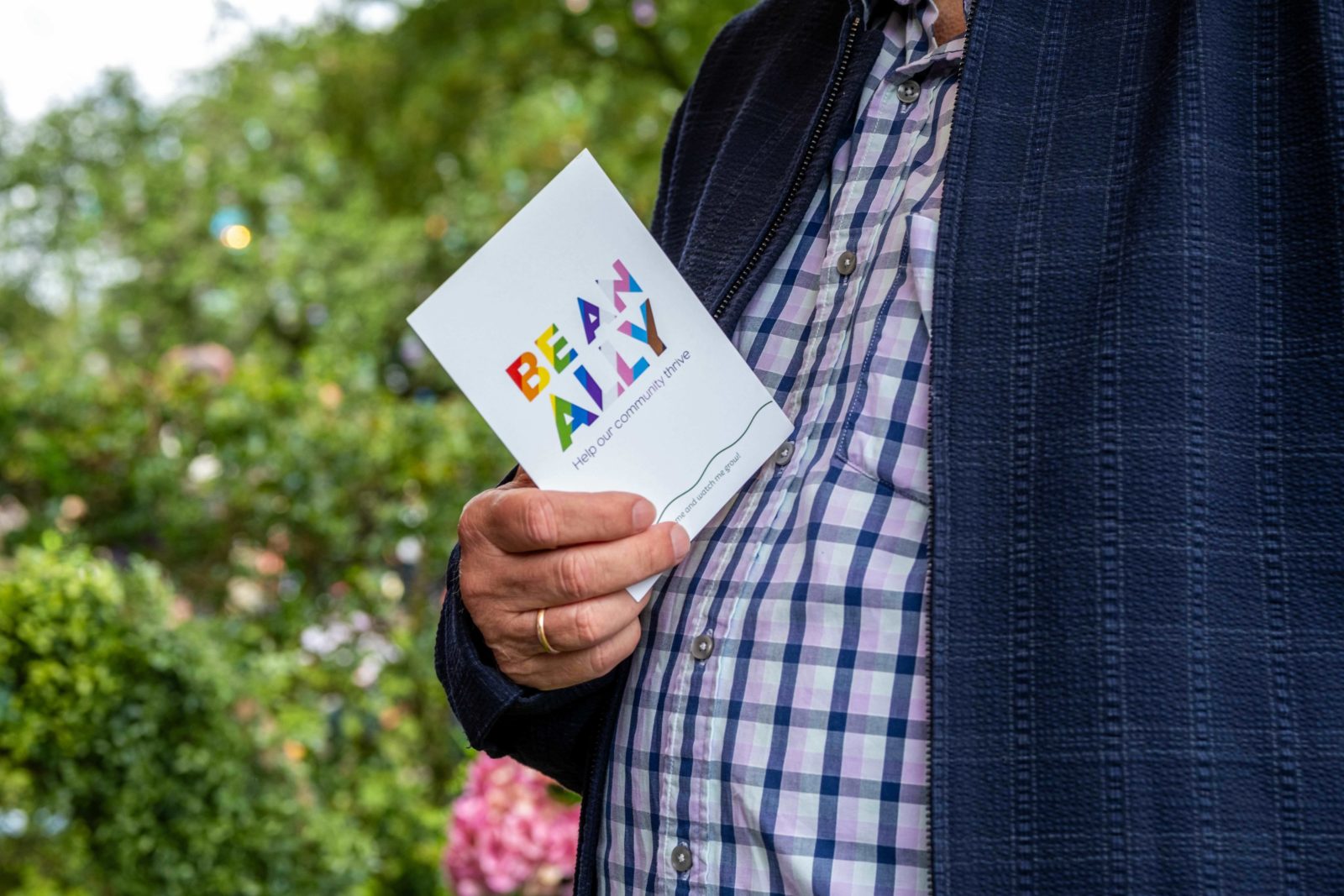 A man's torso is photographed, in his hand is a 'Be an Ally' postcard. He wears a checkered shirt and is in the park.