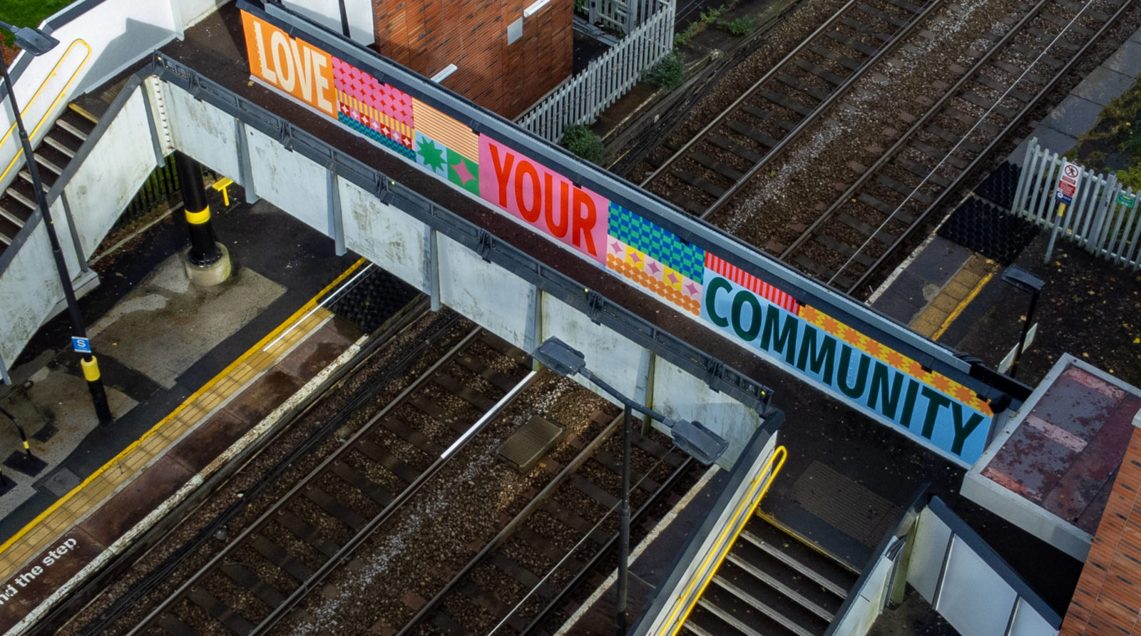The mural is photographed at an arial view, it reads 'Love Your Community', surrounded by pattern and is painted on a train station bridge.