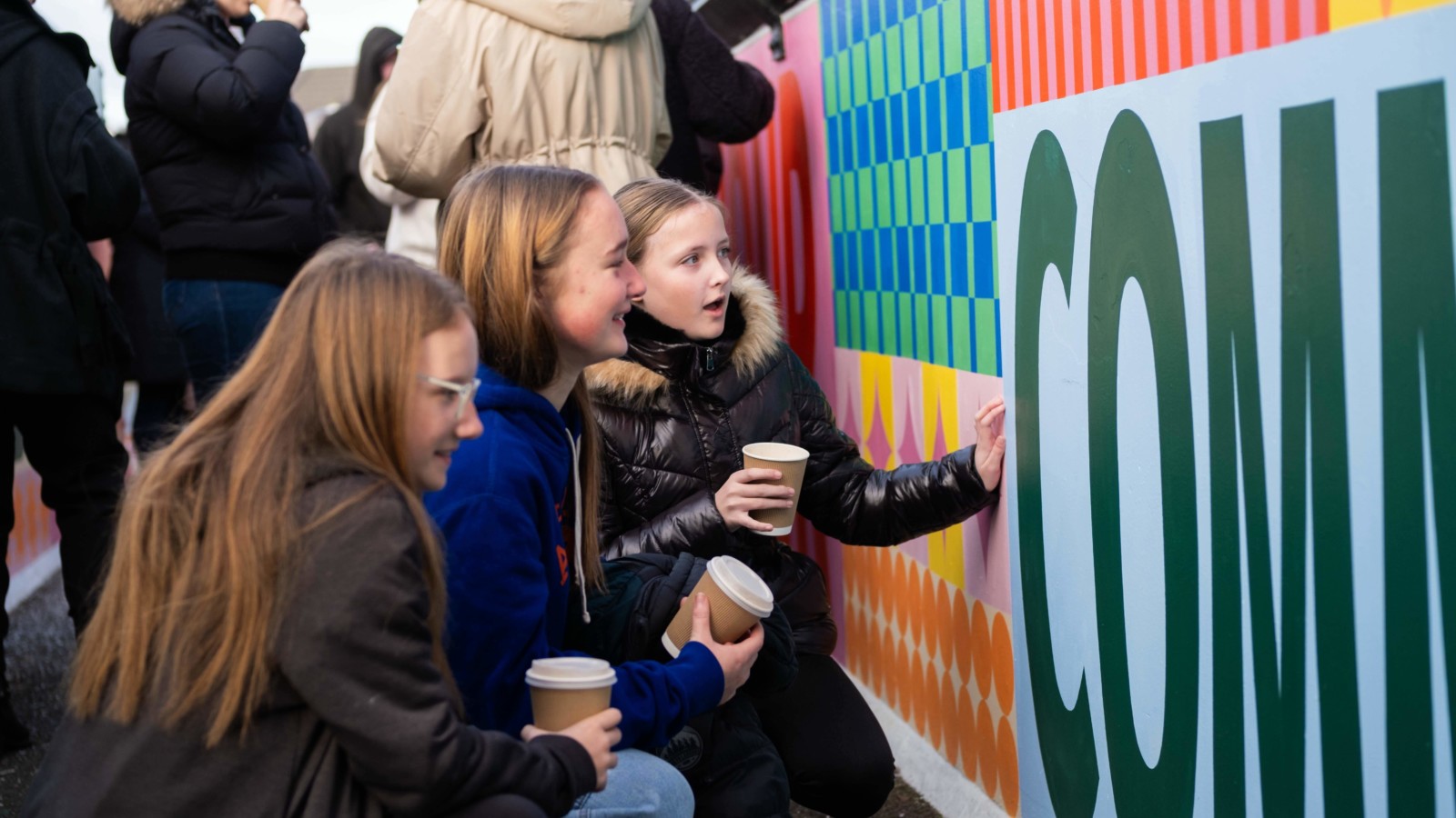 3 young people holding hot drinks look closely at the mural, one is talking with one hand on the wall and the other 2 are smiling.