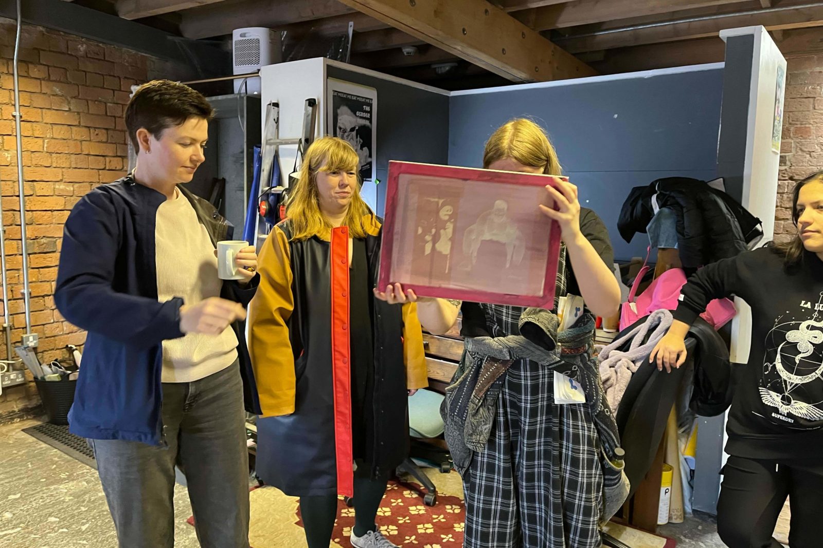 4 people stand in a print room, one is holding up a mesh screen with designs on it and looking at them closely. Another stands with a mug in one hand and others also look at the screen from afar.