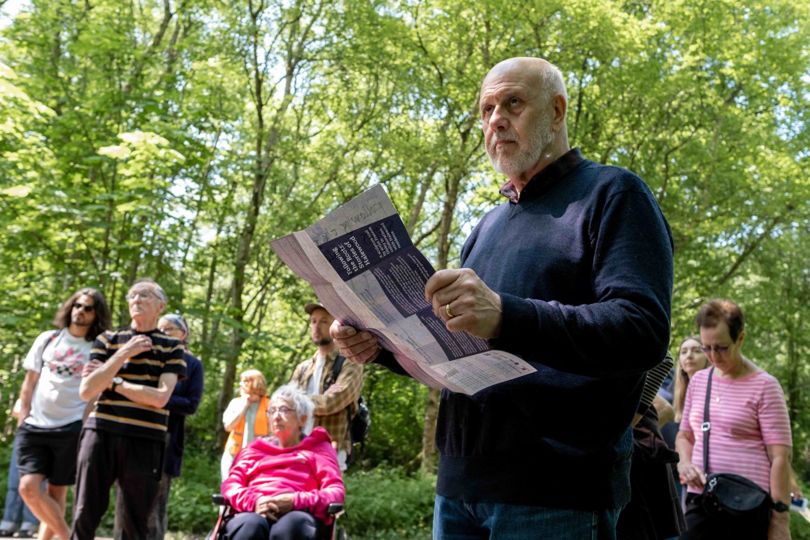 A man is holding a map in the forest, looking up. Several other people are in the background looking in the same direction.