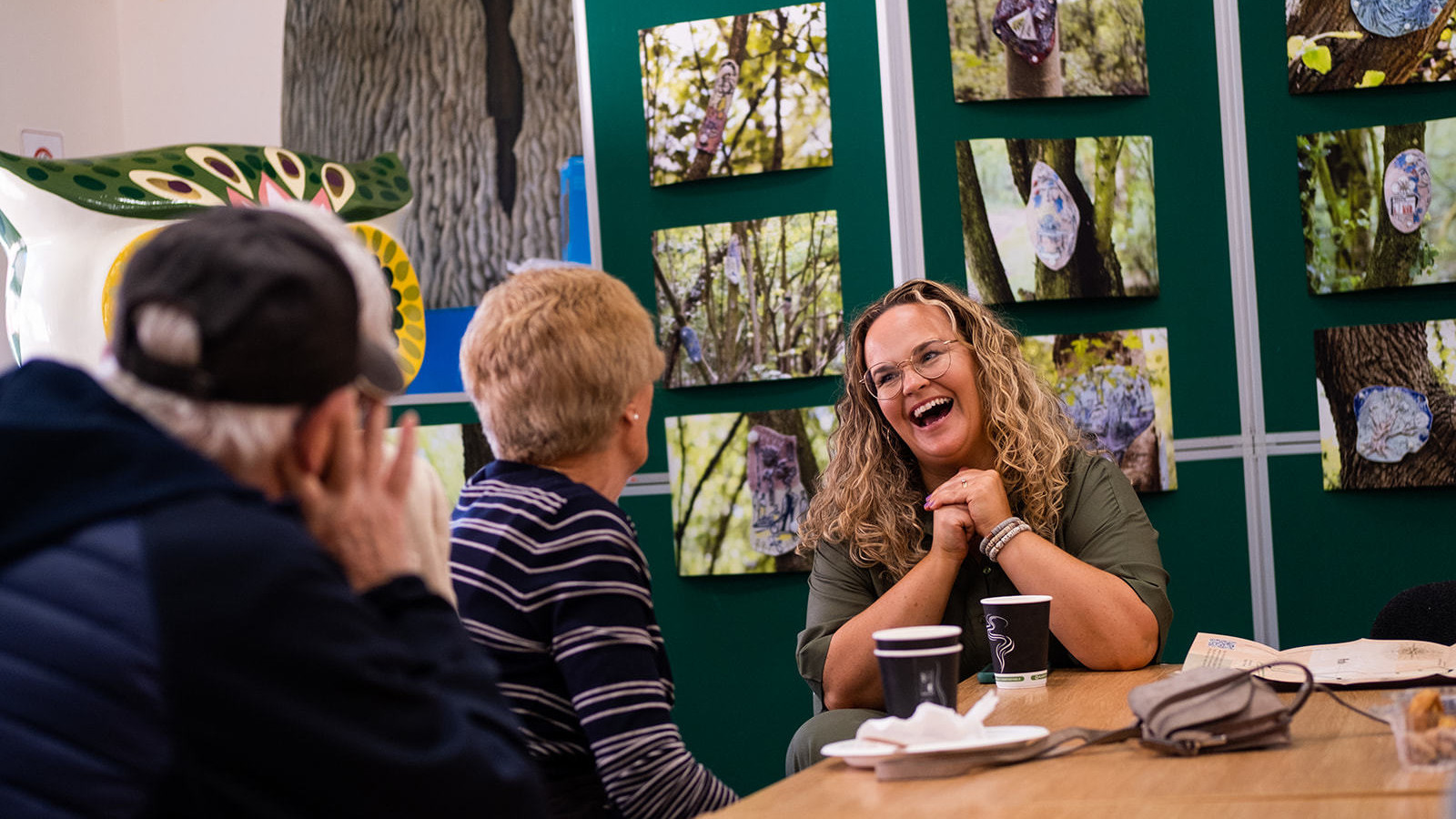A woman with blonde curly hair and round glasses laughs whilst in conversation with an elderly couple. Behind them are photographs of Frances Disley’s sculpture trail ‘Following the Roots’ in Halewood Park.