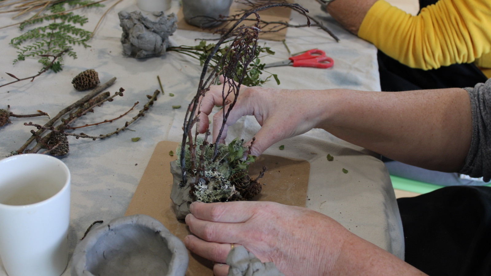 A pair of hands are photographed at a table moulding clay around some twigs and other natural objects.