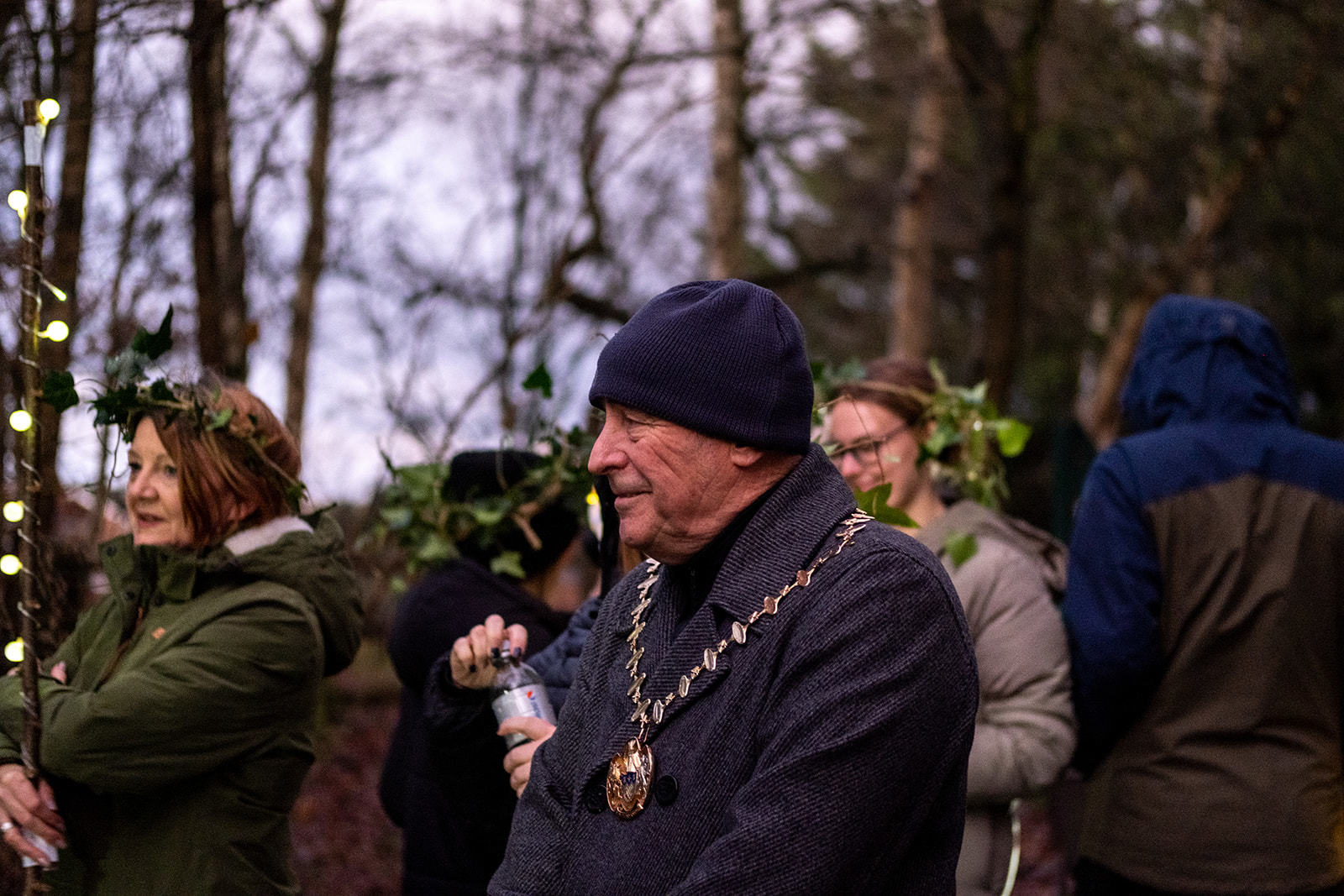 The mayor of Halewood smiling, behind him people are wearing headrests made of leaves, one is carrying a stick wrapped with lights.