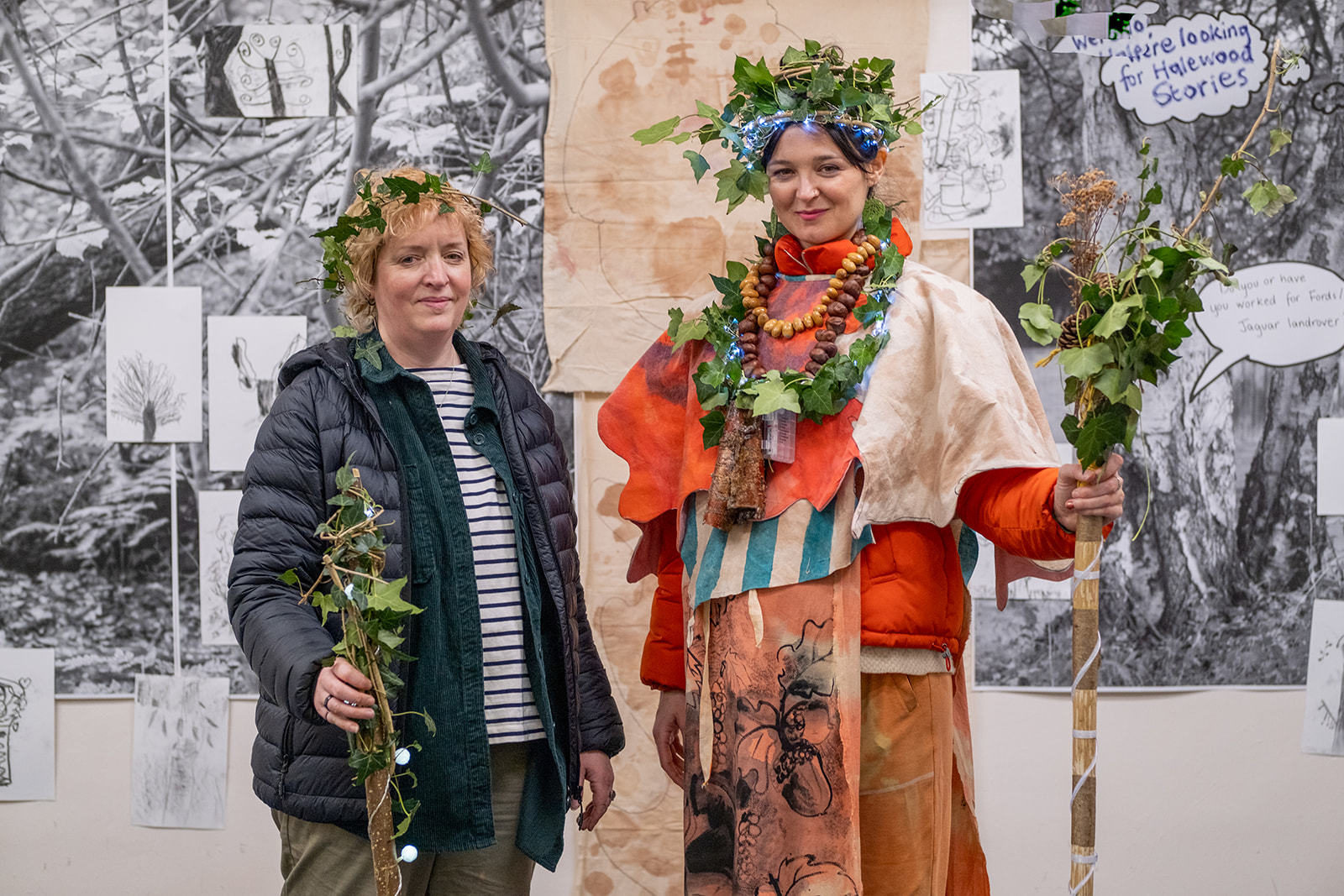 Frances Disley stood with performer Maria Malone, who is holding a staff wrapped in leaves, and a headless made of leaves and lights, along with printed fabrics with illustrations of elements of the woods on them.